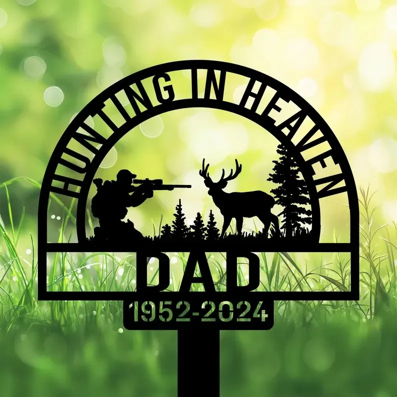 Black yard stake sign reading "Hunting in Heaven Dad 1952-2024," with hunter, deer, and pine silhouettes in grass