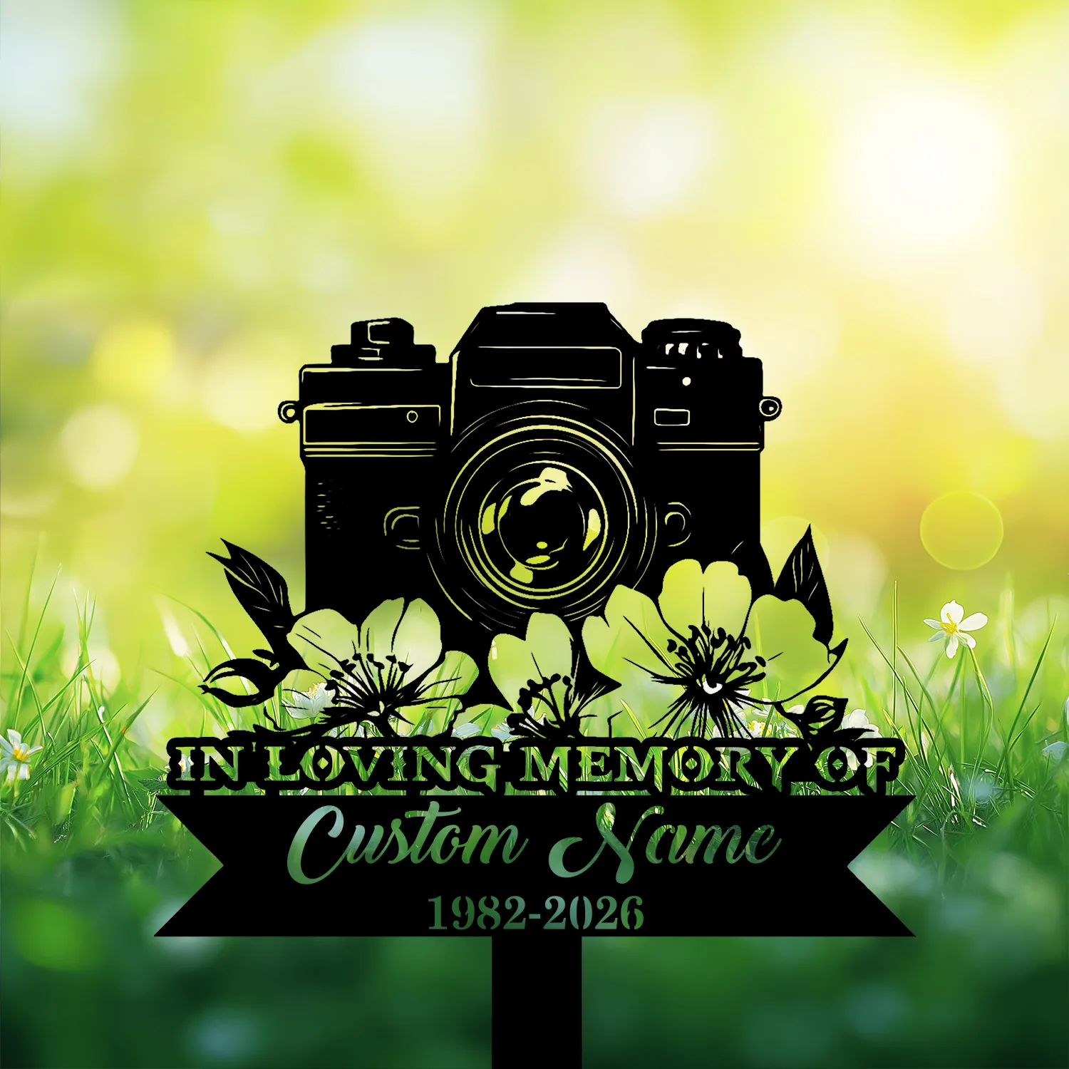 Black silhouette memorial sign with a camera and flowers reading "In Loving Memory" and placeholder "Custom Name 1982-2026" against a sunlit grassy background