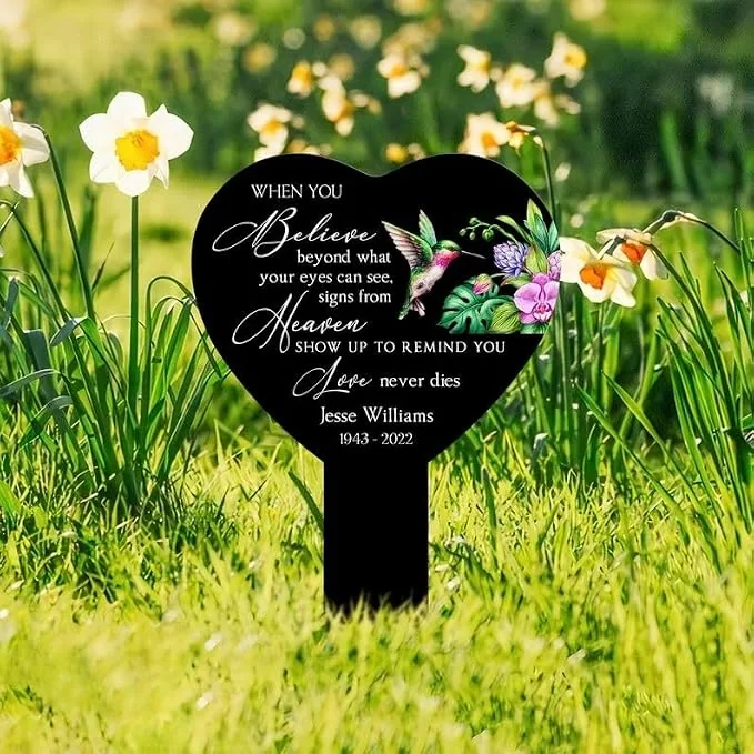 Heart-shaped black memorial stake in grassy garden among daffodils reading: “When you believe beyond what your eyes can see, signs from Heaven show up to remind you love never dies” with name Jesse Williams 1943–2022 and hummingbird floral design.