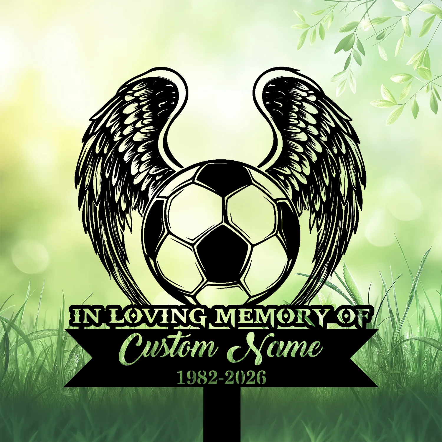 Black silhouette memorial sign with a soccer ball and angel wings over grass and leafy green bokeh background, text reading "IN LOVING MEMORY" and placeholder "Custom Name 1982-2026"
