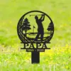 Black metal memorial garden sign showing a golfer silhouette swinging inside a circular golf course scene with trees, the word PAPA and dates 1944 - 2025 staked in grassy field with yellow flowers