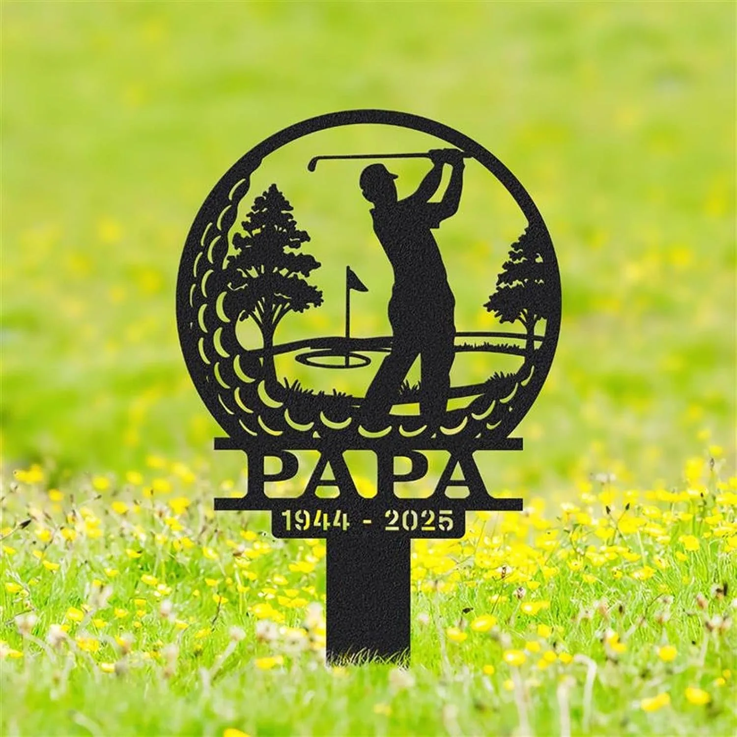 Black metal memorial garden sign showing a golfer silhouette swinging inside a circular golf course scene with trees, the word PAPA and dates 1944 - 2025 staked in grassy field with yellow flowers