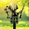 Black metal memorial sign reading "DAD 1932-2025" with deer heads, compound bow and rifle on a stake set in a sunlit grassy yard