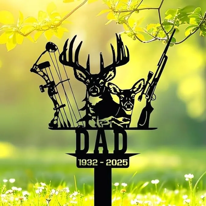 Black metal memorial sign reading "DAD 1932-2025" with deer heads, compound bow and rifle on a stake set in a sunlit grassy yard