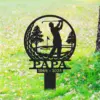 Black metal memorial yard sign showing a golfer silhouette swinging inside a circular frame with trees and a flag, text 'PAPA' and dates '1944 - 2025' on green lawn