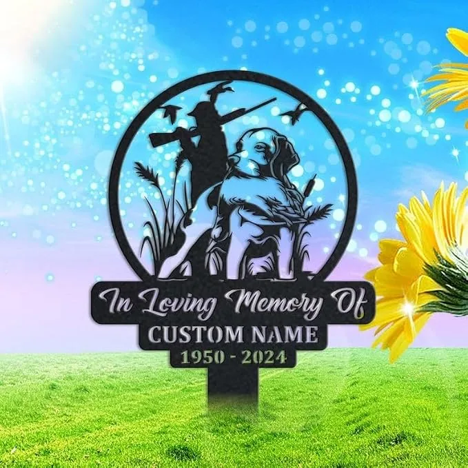 Circular black metal memorial sign with a hunter, dog, reeds and flying ducks, text reading "In Loving Memory Of" with placeholder "CUSTOM NAME" and dates "1950 - 2024", displayed on grassy lawn with blue sky and sunflowers.