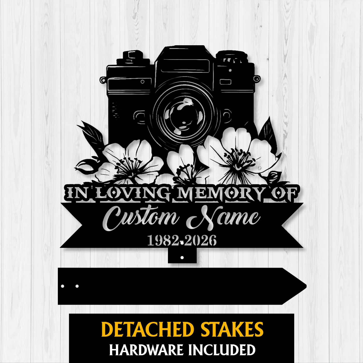 Black metal memorial sign shaped like a camera with flowers, reading "IN LOVING MEMORY OF" and a cursive placeholder "Custom Name" with dates 1982-2026, on a light wood background