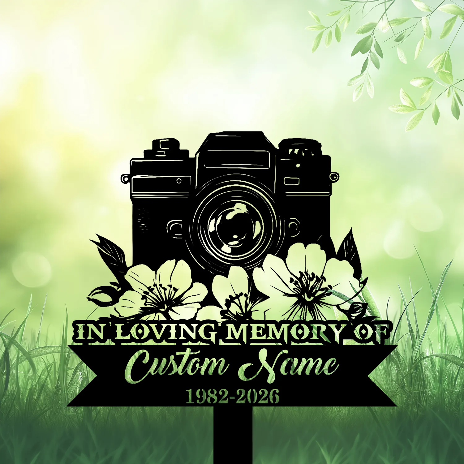 Black silhouette memorial sign with a vintage camera above flowers, ribbon reading "IN LOVING MEMORY OF" and placeholder "Custom Name 1982-2026" on green grassy background