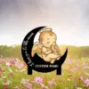 Metal garden sign showing a sleeping angel baby with halo perched on a black crescent moon reading "I love you to the moon" and placeholder text "CUSTOM NAME" against a wildflower field