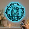 LED wall art of two embracing skeletons inside a floral round frame with names "LIAM & EMMA", glowing blue on a living room wall above a mantel with flowers, candle, photo, and remote control