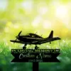 Black silhouette memorial sign of a small airplane over green grass and white flowers with text reading "IN LOVING MEMORY OF Custom Name 1982-2026" against a sunlit bokeh background