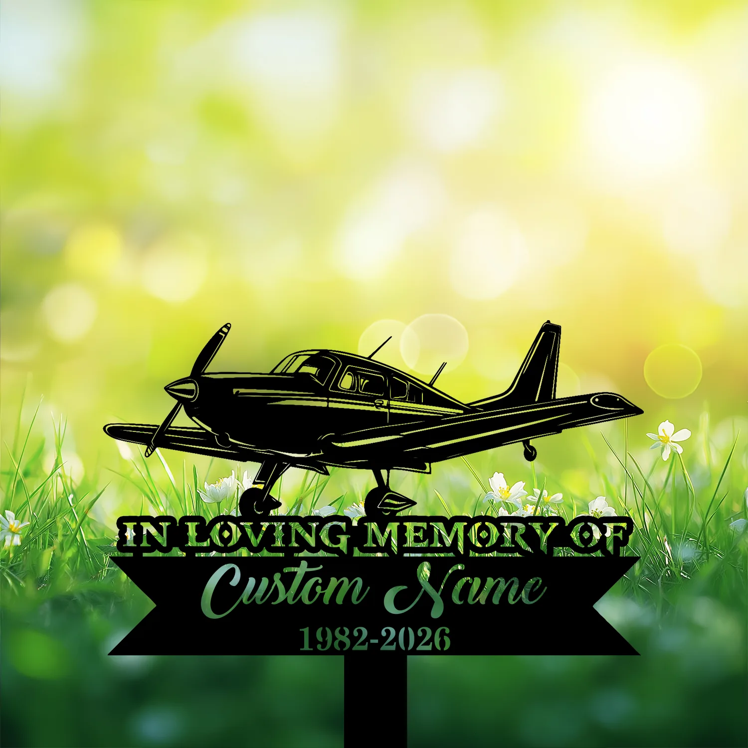 Black silhouette memorial sign of a small airplane over green grass and white flowers with text reading "IN LOVING MEMORY OF Custom Name 1982-2026" against a sunlit bokeh background