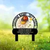 Black memorial stake with custom name and dates encircling a daisy and ladybug illustration, placed in a sunny grassy field with small yellow flowers