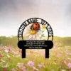 Memorial garden sign mockup with a daisy and ladybug illustration, customizable name and years arched above, and a condolence inscription on a bench silhouette in a wildflower field