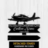 Black metal memorial sign shaped like a small propeller airplane mounted on a light wood background, text reads "In Loving Memory of Custom Name 1982-2026" with detached stakes shown below