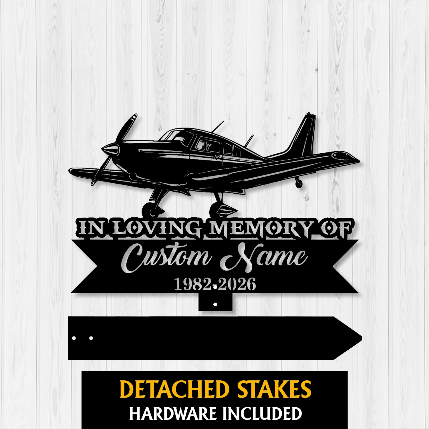 Black metal memorial sign shaped like a small propeller airplane mounted on a light wood background, text reads "In Loving Memory of Custom Name 1982-2026" with detached stakes shown below