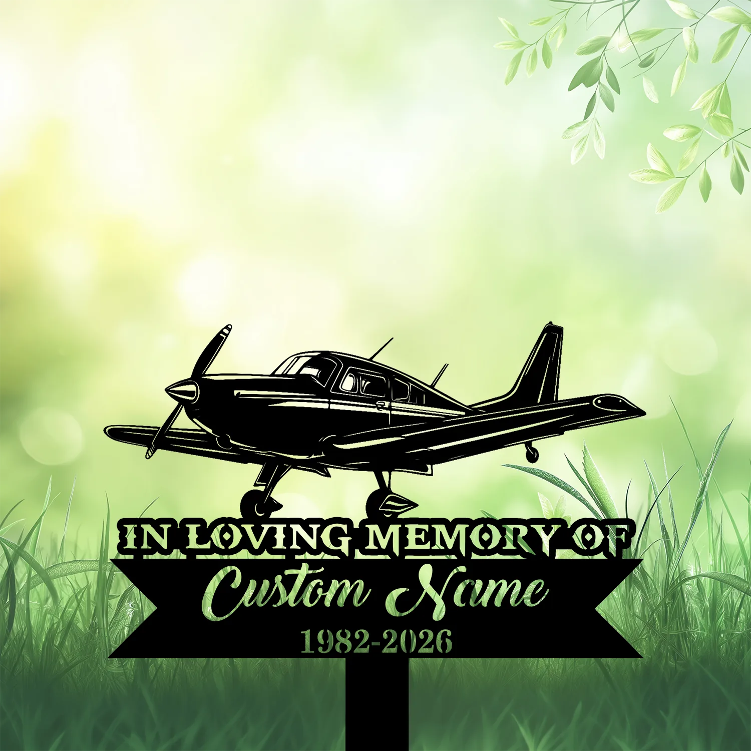 Black silhouette of a small single‑engine airplane above a memorial sign reading "IN LOVING MEMORY" and "Custom Name 1982-2026" against a soft green bokeh background with grass and leaves