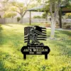 Memorial yard sign in a sunny park showing combat boots, helmet, and American flag with text "In Loving Memory of Jack William 1956 - 2025" placed in grassy area near picnic shelter