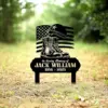 Black metal memorial yard sign with combat boots, helmet and American flag reading "In Loving Memory of Jack William 1956 - 2025" placed in a grassy outdoor setting