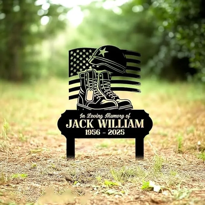 Black metal memorial yard sign with combat boots, helmet and American flag reading "In Loving Memory of Jack William 1956 - 2025" placed in a grassy outdoor setting
