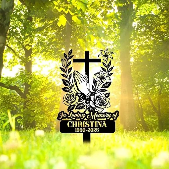 Black memorial cross sign in a sunlit forest clearing reading "In Loving Memory of CHRISTINA 1980-2025" with praying hands and floral illustrations