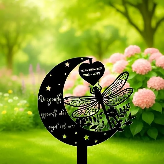 Black metal memorial garden stake in crescent moon shape with dragonfly cutout, text 'Dragonfly appears when angel is near' and heart plaque reading 'Bella Thompson 1993 - 2025', placed in a green garden near pink hydrangeas