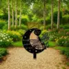 Black crescent garden memorial stake with dragonfly cutout and engraved name, placed on a gravel path amid flowering garden beds and birch trees