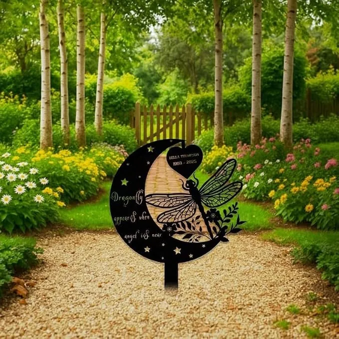 Black crescent garden memorial stake with dragonfly cutout and engraved name, placed on a gravel path amid flowering garden beds and birch trees