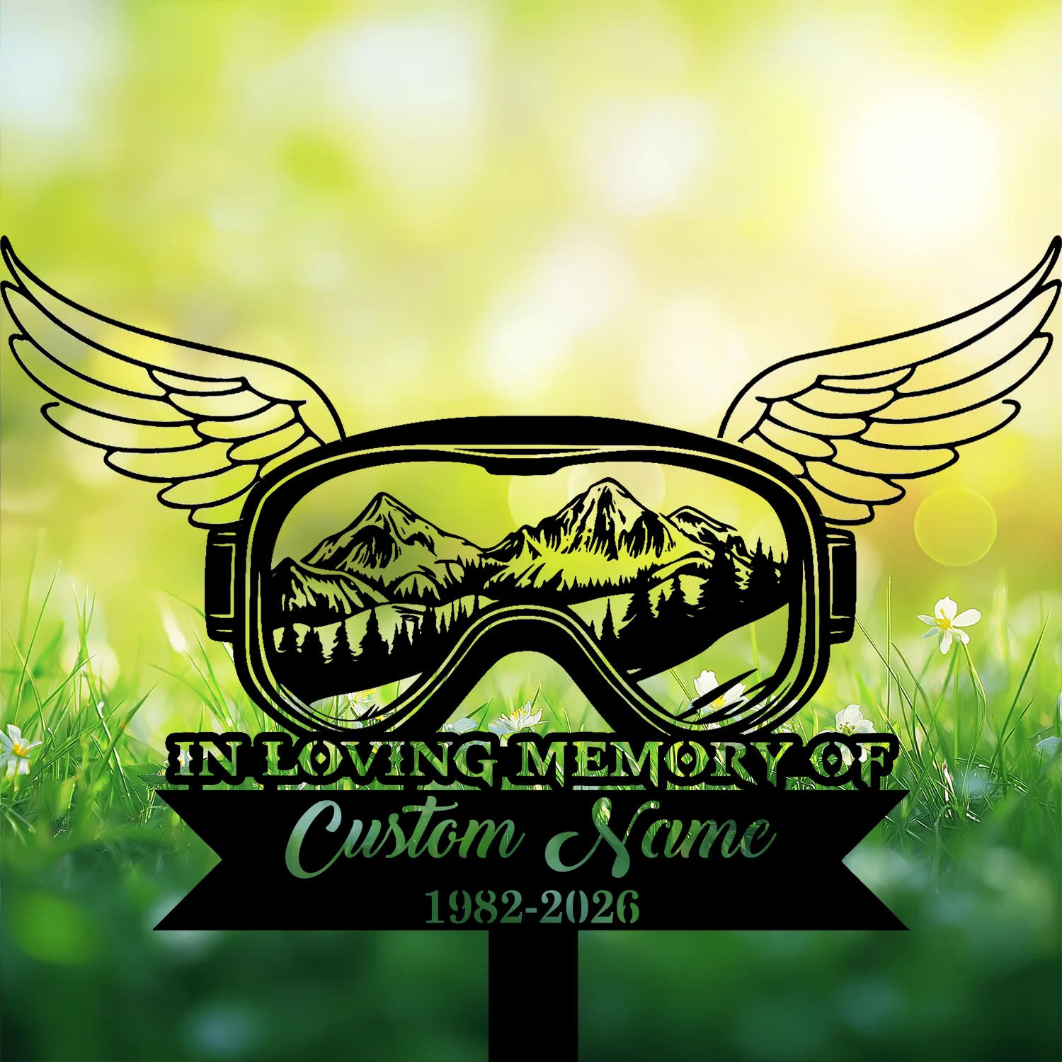 7-4 Black memorial sign shaped like winged goggles with mountain scene inside, text 'IN LOVING MEMORY OF', placeholder 'Custom Name' and dates '1982-2026' against sunny grassy background