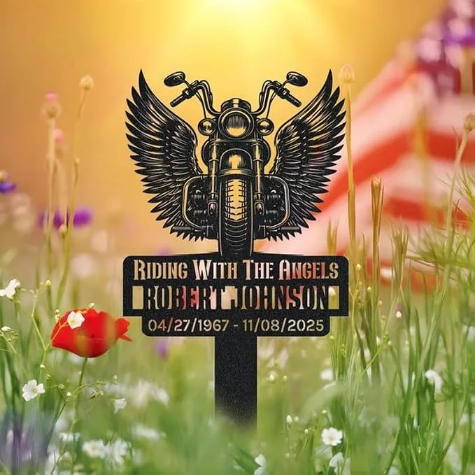 Memorial metal stake in a flower meadow with a winged motorcycle design and text “Riding With The Angels” and a name and dates beneath