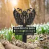 Black metal grave marker shaped like a winged motorcycle with text 'Riding With The Angels' and name 'Robert Johnson' plus dates, placed among spring flowers in a forest floor