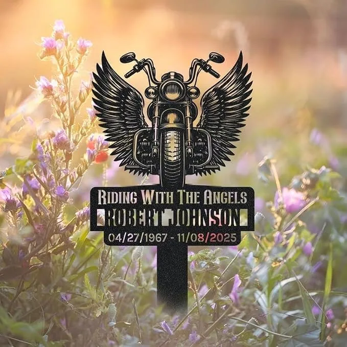 Black metal memorial sign with a winged motorcycle and text “Riding With The Angels Robert Johnson 04/27/1967 - 11/08/2025” placed among sunlit wildflowers