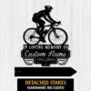 Black metal memorial sign showing a cyclist silhouette above text reading "In Loving Memory of Custom Name 1987-2026" on a pale wood background