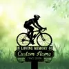 Silhouette of a cyclist on a road bike above a memorial plaque reading "IN LOVING MEMORY OF" and placeholder "Custom Name 1987-2026" on a grassy green background