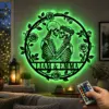 Round metal wall art of two embracing skeletons with roses and the names "LIAM & EMMA" backlit in green, hung above a living room shelf with candles and a hand holding an LED remote