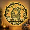 Backlit round wall art of two embracing skeletons with floral wreath and 'LIAM & EMMA' text, LED-lit on a textured wall next to candles and a remote