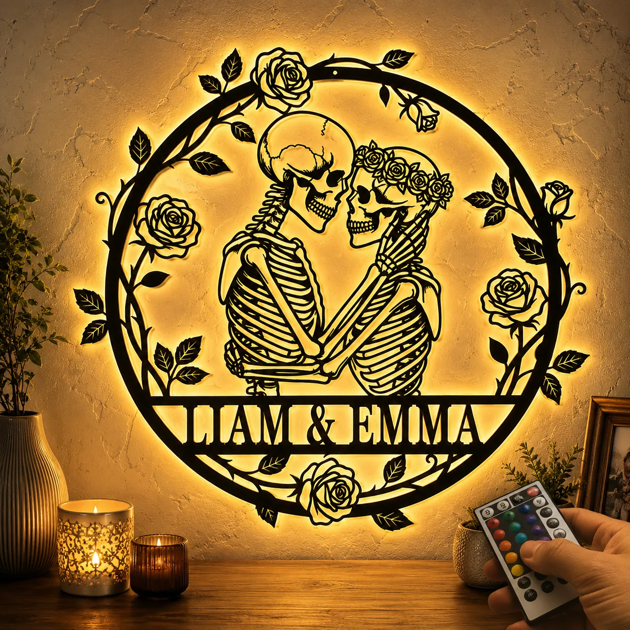 Backlit round wall art of two embracing skeletons with floral wreath and 'LIAM & EMMA' text, LED-lit on a textured wall next to candles and a remote