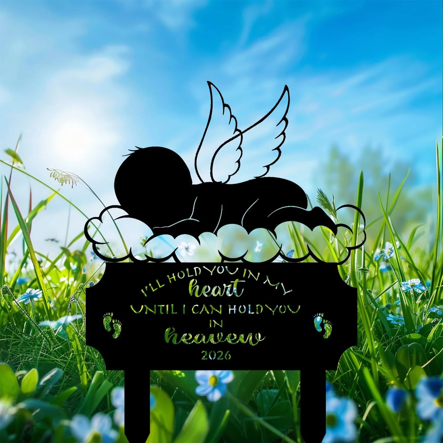 Black silhouette lawn memorial sign showing a sleeping baby with angel wings on a cloud, text cutout reading "I'll hold you in my heart until I can hold you in heaven 2026", set in grassy field with blue sky and wildflowers