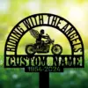 Angel-Biker-1 Black metal memorial sign showing a winged motorcycle rider silhouette under arched text 'RIDING WITH THE ANGELS' and a customizable name with dates '1954-2024' against a sunlit grassy background