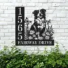 Black metal house sign mounted on a white brick wall showing numbers 1565 and text "Fairway Drive" with a dog, flowers and butterflies, greenery below