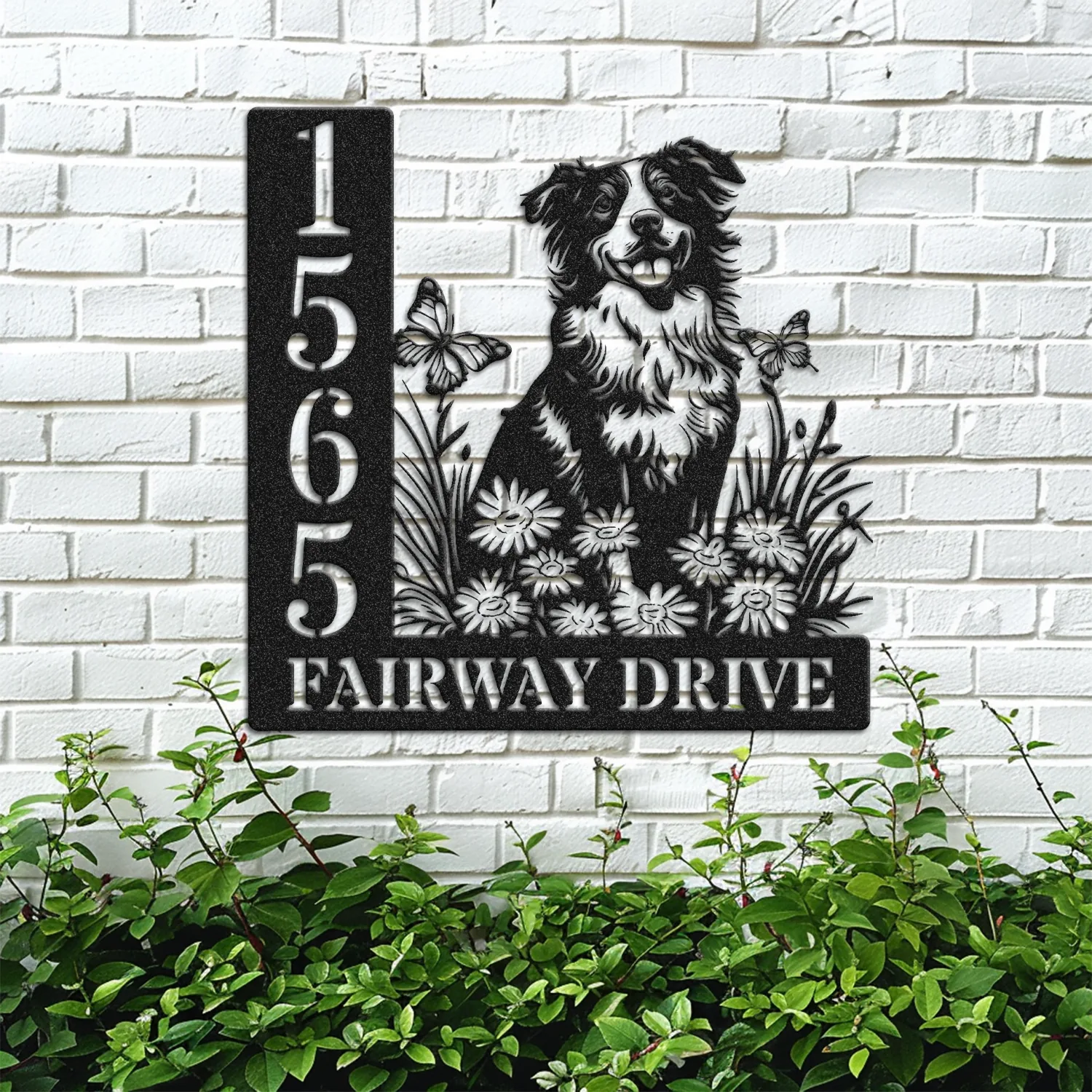 Black metal house sign mounted on a white brick wall showing numbers 1565 and text "Fairway Drive" with a dog, flowers and butterflies, greenery below
