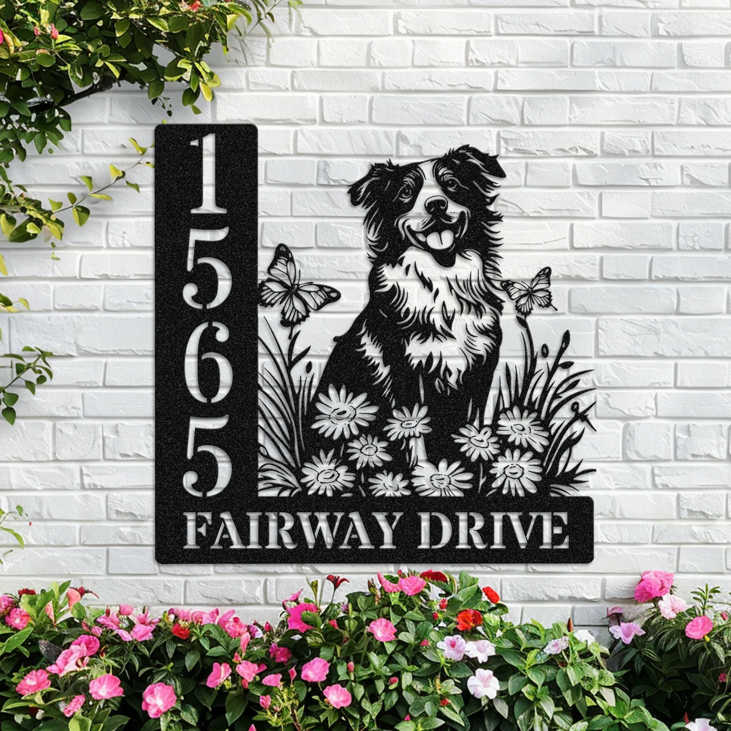 Black metal address sign mounted on white brick wall showing numbers 1565 and text FAIRWAY DRIVE with cutout dog, flowers and butterflies, pink flowers below