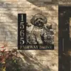 Black metal house sign on a light brick wall showing numbers 1565 and text FAIRWAY DRIVE with a dog illustration surrounded by flowers and butterflies