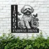 Black metal house sign mounted on a white brick wall showing address number 1565 and the text FAIRWAY DRIVE with a happy fluffy dog surrounded by flowers and butterflies above garden plants