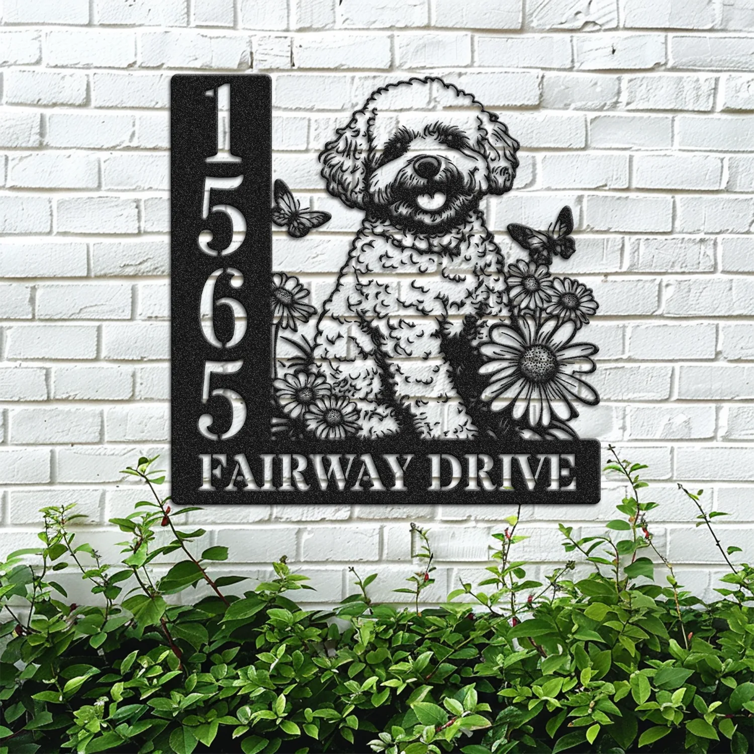 Black metal house sign mounted on a white brick wall showing address number 1565 and the text FAIRWAY DRIVE with a happy fluffy dog surrounded by flowers and butterflies above garden plants