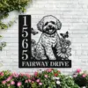 Black metal house sign mounted on a white brick wall showing numbers 1565 and the text 'FAIRWAY DRIVE' with a cutout illustration of a fluffy dog, daisies, butterflies and surrounding pink flowers at the base