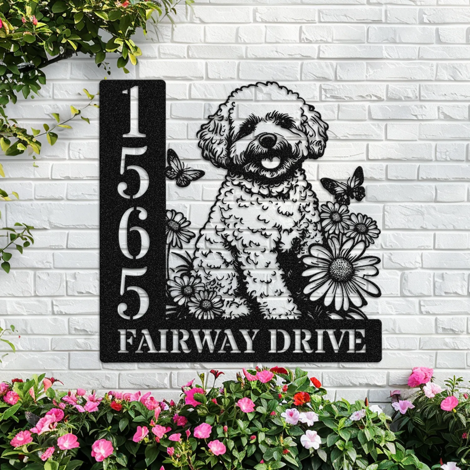 Black metal house sign mounted on a white brick wall showing numbers 1565 and the text 'FAIRWAY DRIVE' with a cutout illustration of a fluffy dog, daisies, butterflies and surrounding pink flowers at the base