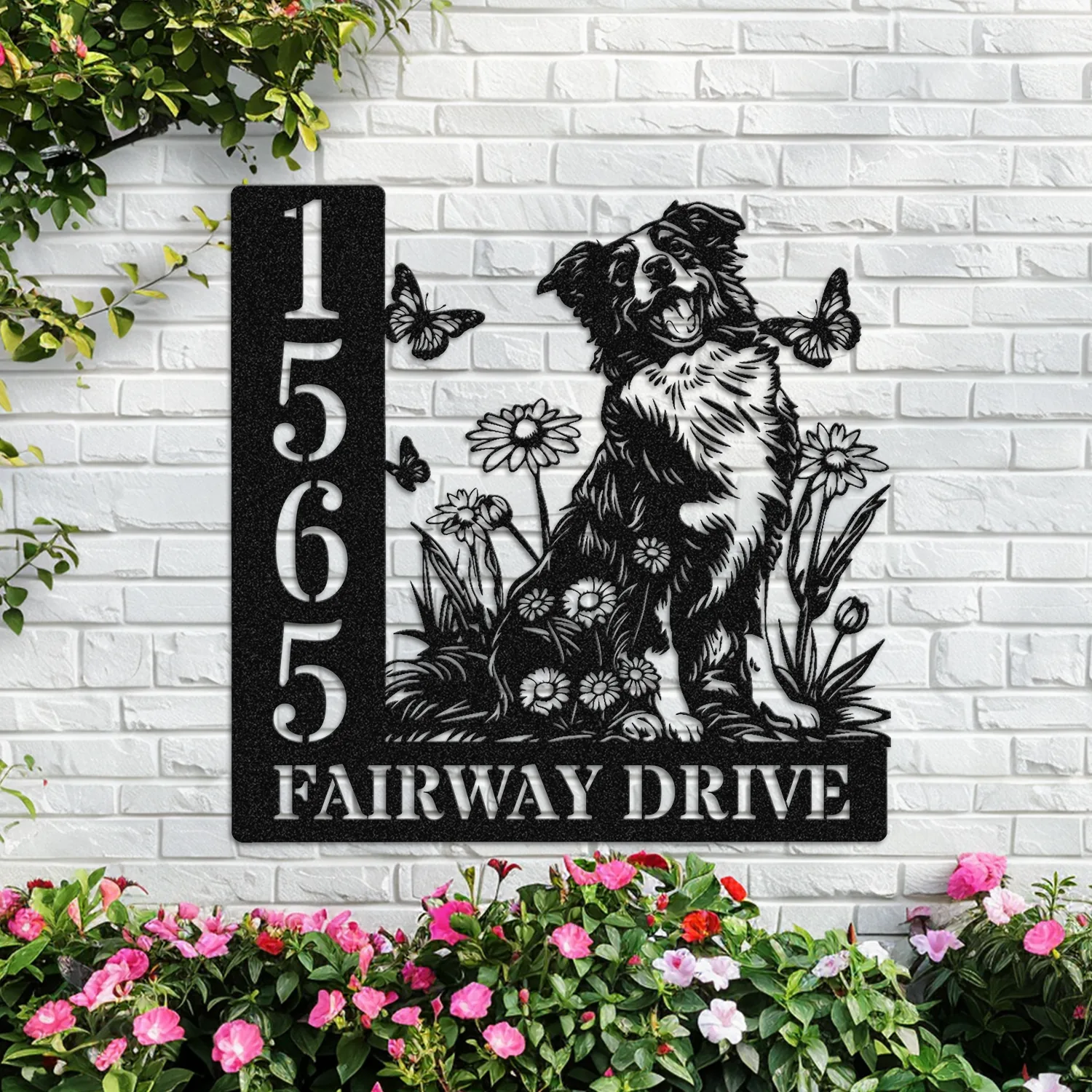 Border-Collie-Sitting-with-Butterfly-4 Black metal house sign on a white brick wall showing address numbers 1565 and text 'FAIRWAY DRIVE' with a dog, butterflies and flowers, surrounded by pink flowering plants
