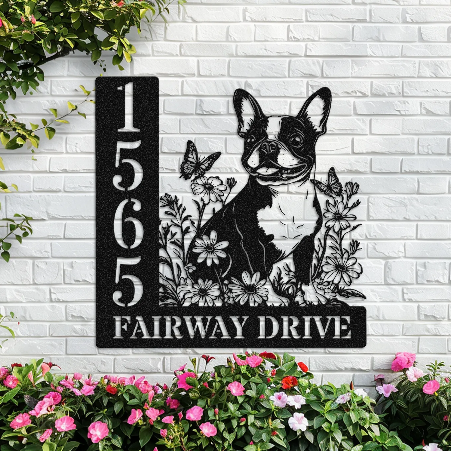 Boston-Terrier-Sitting-with-Butterfly-4 Black metal house sign mounted on a white brick wall showing address 1565 and text "FAIRWAY DRIVE" with a decorative Boston Terrier, flowers and butterflies, pink flower bed below