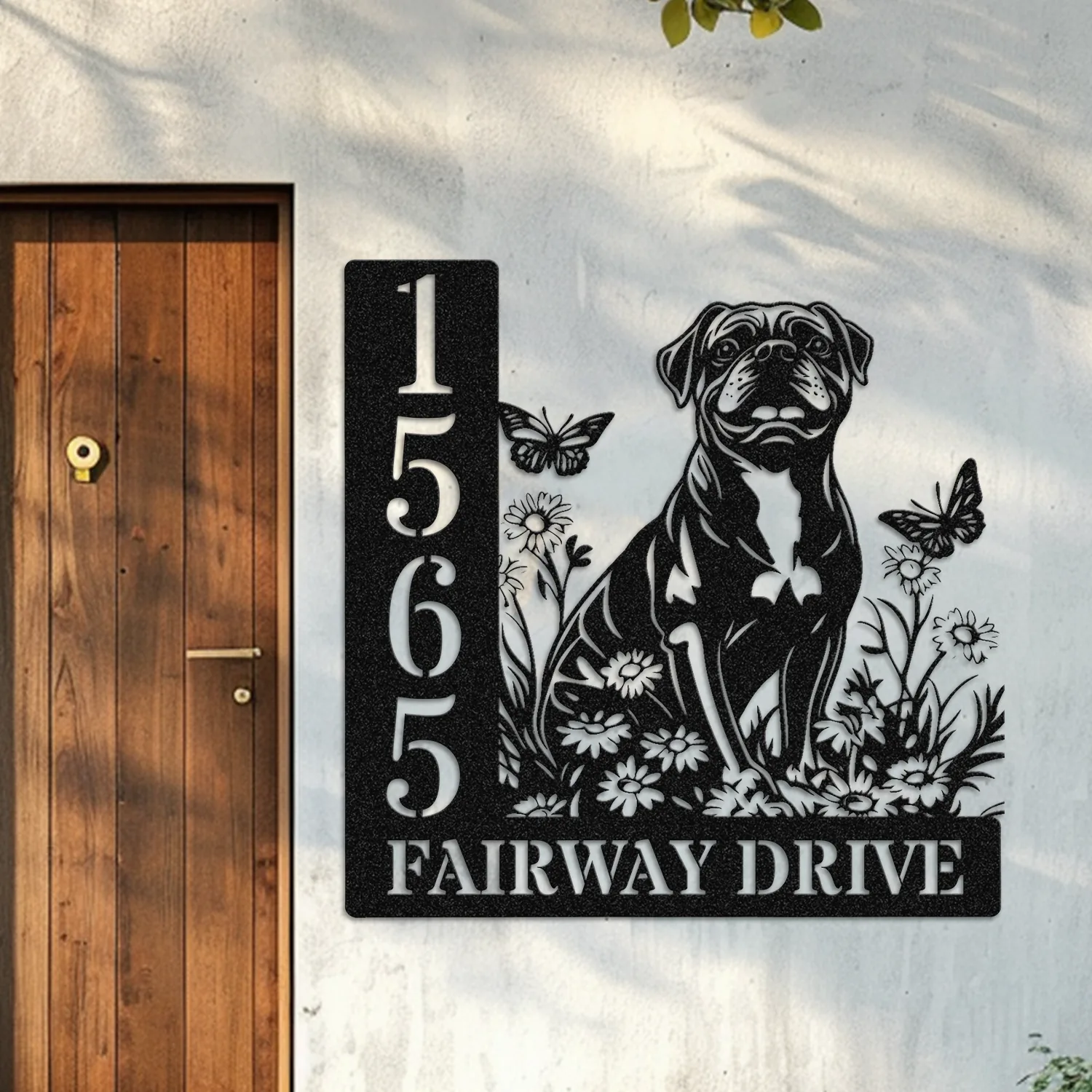Black metal house sign mounted next to a wooden door showing numbers 1565 and text "Fairway Drive" with a decorative dog, flowers, and butterflies on a white stucco wall